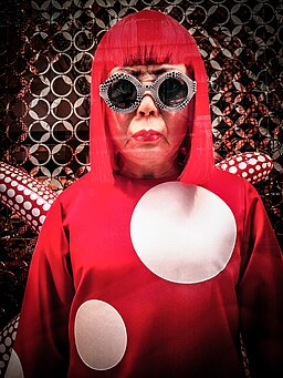 Foto frontal de Yayoi Kusama usando óculos escuros e vestido vermelho com polka dots brancos, característicos de sua arte.