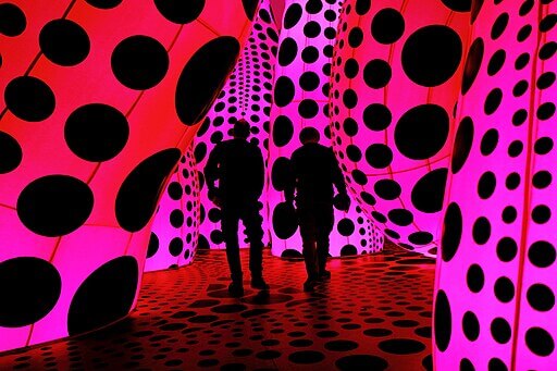 Duas silhuetas humanas caminham em meio à instalação imersiva de Yayoi Kusama.